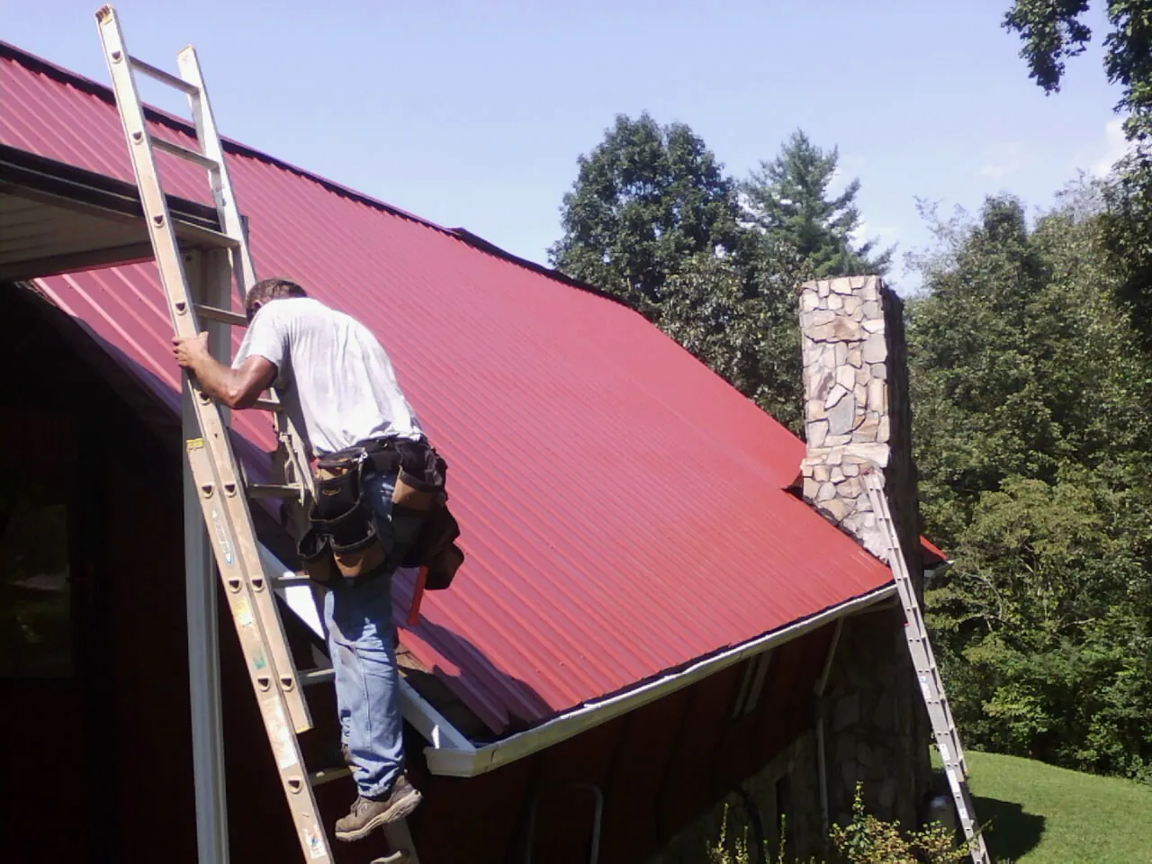 Roofer on a ladder beside a red metal roof and stone chimney — Soffit & Fascia Repair by Roofing Brothers in Emmaus